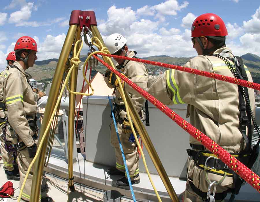 Firefighters conducting high-angle rescue training, using red, yellow, and blue safety ropes rigged to a tripod for secure fall protection