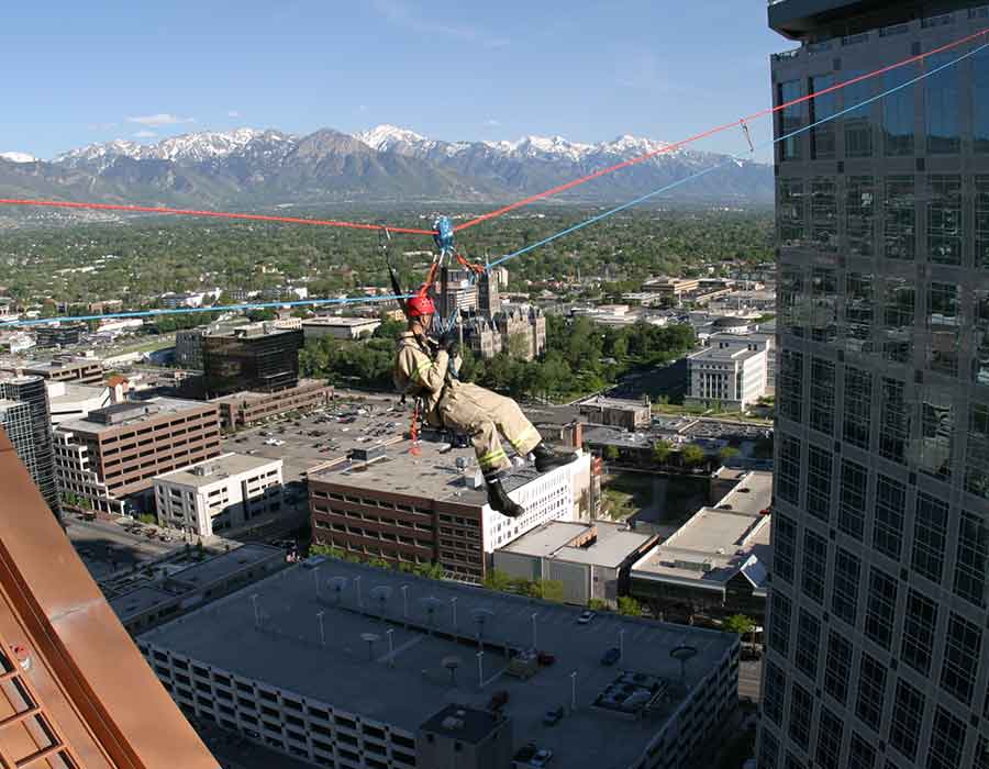 Firefighter suspended on a high-angle rescue rope system between buildings, using red and blue safety ropes for fall protection